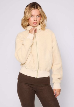 Beige, fuzzy, ritssluiting jack met een hoge kraag, geribde manchetten en zoom, voorzien van een lichtgroene ritssluiting. Draagt over een bruin rok.