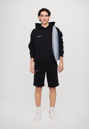 Black hooded sweatshirt with a denim accent, paired with black shorts. Notable features include a logo print and a relaxed fit. White sneakers.