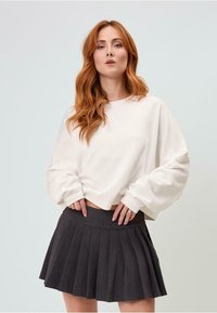 Sudadera corta blanca con mangas largas, combinada con una minifalda oscura ajustada y plisada, que muestra una textura suave y un diseño relajado.