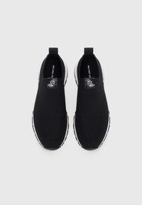 Chaussures noires à enfiler avec une tige en mesh, des semelles en caoutchouc blanches et un logo circulaire sur le dessus. Texture lisse et design épuré.