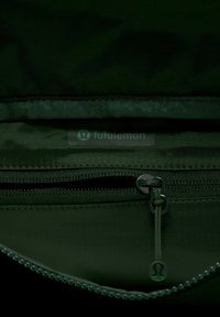 Ciemnozielona saszetka na zamek błyskawiczny ma gładką teksturę, z logo i uchwytem do zamka w dopasowanych ciemnych odcieniach. Wnętrze jest funkcjonalne i kompaktowe.