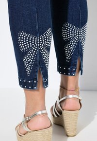 Tmavě modré džíny capri s velkými kamínkovými mašlemi na zadní straně, s bočními rozparky a dekorativními cvočky na manžetách.