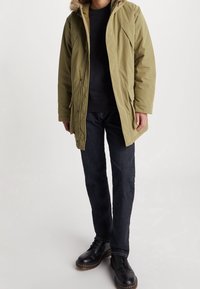 Parka verde oliva con capucha forrada de piel, con una cremallera frontal, dos bolsillos laterales y un ajuste holgado, combinada con vaqueros oscuros y botas negras.