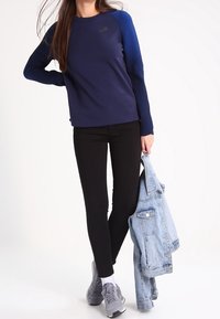 Mörkblå långärmad atletisk topp med ribbade detaljer på axlarna, tillsammans med svarta leggings och grå sneakers, hållande en ljusblå denimjacka.