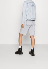 Giacca di jeans azzurro chiaro con logo sul retro, abbinata a pantaloni shorts in denim grigi e stivaletti Chelsea in pelle nera. Calze bianche visibili.