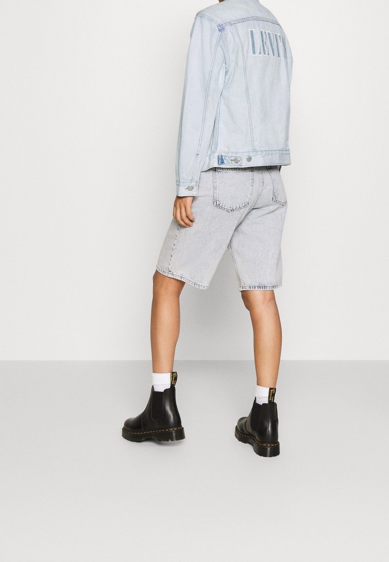 Giacca di jeans azzurro chiaro con logo sul retro, abbinata a pantaloni shorts in denim grigi e stivaletti Chelsea in pelle nera. Calze bianche visibili.