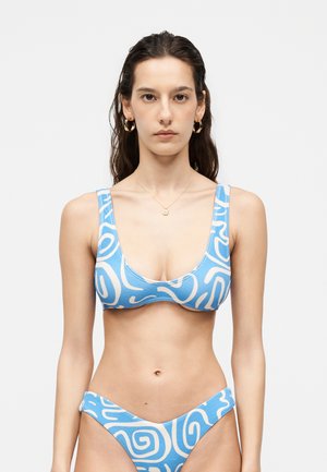 Kvinde stående iført en bikini med blå og hvide abstrakte mønstre, gyldne hoops øreringe og en lille vedhæng halskæde.