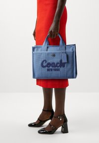 Niebieska torba płócienna z haftowanym logo "Coach New York", mocne uchwyty i zawieszka, w zestawieniu z czerwoną sukienką i czarnymi szpilkami.