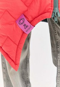 Prošívaná ružová vesta s výraznou fialovou štítkom s nápisom „SEE IT ALL NAOKO“ a štýlizovanou grafikou oka, doplnená o sivé džínsy.