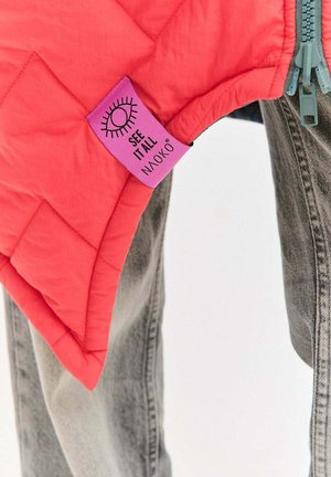 Prošívaná ružová vesta s výraznou fialovou štítkom s nápisom „SEE IT ALL NAOKO“ a štýlizovanou grafikou oka, doplnená o sivé džínsy.