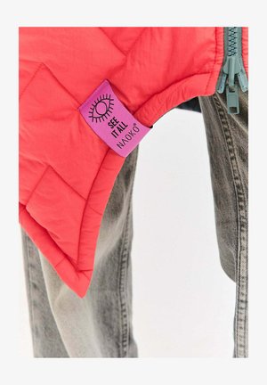 Prošívaná růžová vesta s výraznou fialovou visačkou s nápisem "SEE IT ALL NAOKO" a stylizovaným motivem oka, doplněná o šedé džíny.