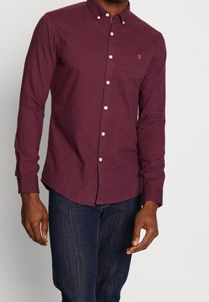 Mand iført en langærmet burgunderfarvet skjorte med knapper og mørkeblå jeans, stående mod en ensfarvet lys baggrund.