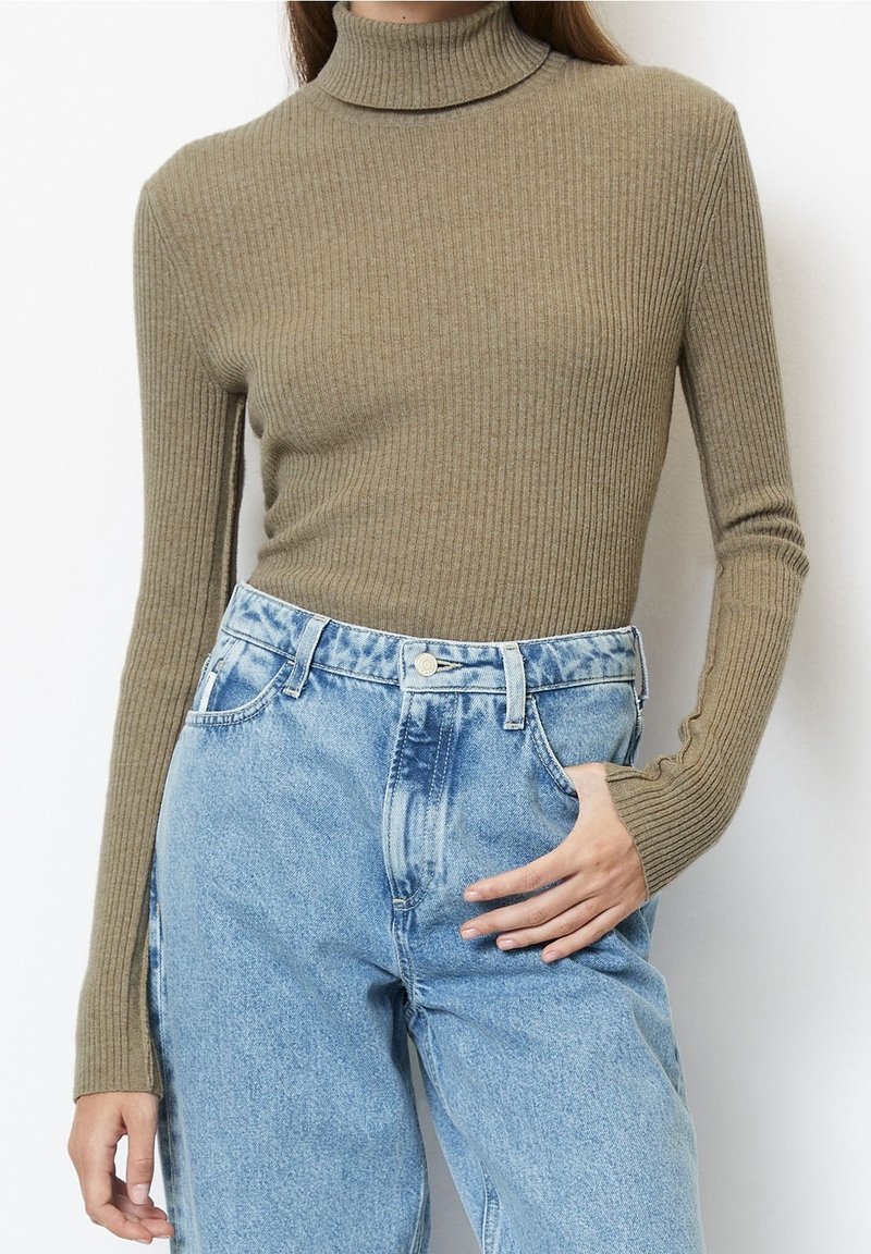 Femme portant un pull à col roulé beige côtelé ajusté, rentré dans un jean denim bleu clair taille haute, une main dans la poche, sur fond blanc.