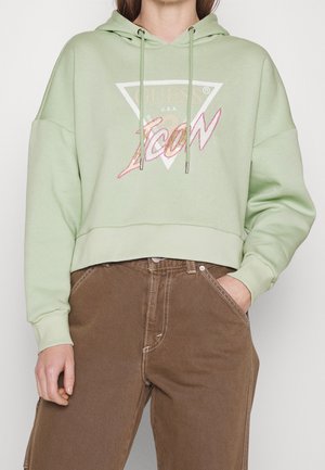 Osoba ubrana w jasnozieloną, krótką bluzę z kapturem z napisem „GUESS Icon” oraz brązowe spodnie z wysokim stanem, z jedną ręką w kieszeni, na białym tle.