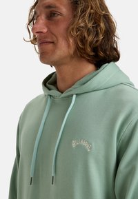 Mint green hoodie with a drawstring, featuring a subtle embroidered logo. Made of soft fabric with a relaxed, casual fit.