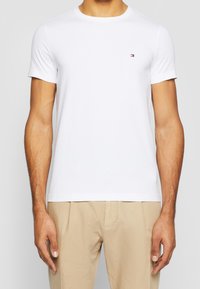 White short-sleeve t-shirt made of cotton, with a crew neck design and a small logo on the chest. Paired with light beige pants.