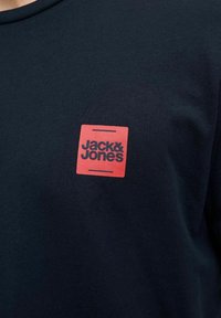 Tumesinine puuvillane T-särk, millel on punane ruudukujuline logo musta tekstiga "Jack & Jones". Ümmargune kaelus ja pehme tekstuur.