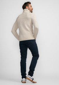 Beige gestrickter Pullover mit hohem Kragen, strukturiertem Vorderteil und geripptem Saum, kombiniert mit dunkelblauen Jeans und lässigen weißen Sneakers.