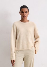 Pull en tricot beige clair avec un col rond, des épaules tombantes et des manches longues. Texture douce. Associé à un pantalon en velours côtelé couleur crème.
