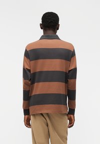 Πόλο μπλούζα με μακριά μανίκια σε καφέ και μαύρες �οριζόντιες ρίγες. Κατασκευασμένη από απαλό ύφασμα, διαθέτει λαιμόκοψη με γιακά και ριμπ μανσέτες.