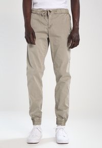 Beige joggerbyxor med dragsko och elastiska muddar, tillverkade av ett lättviktigt tyg. Bärs med vita sneakers, subtil textur och sidofickor.