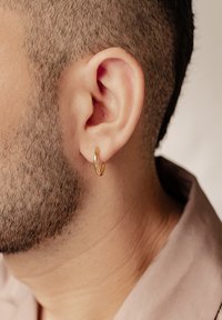 Orecchino a cerchio dorato indossato all'orecchio sinistro, con una forma liscia e arrotondata e una finitura lucida. Primo piano dell'orecchio e dei capelli corti.