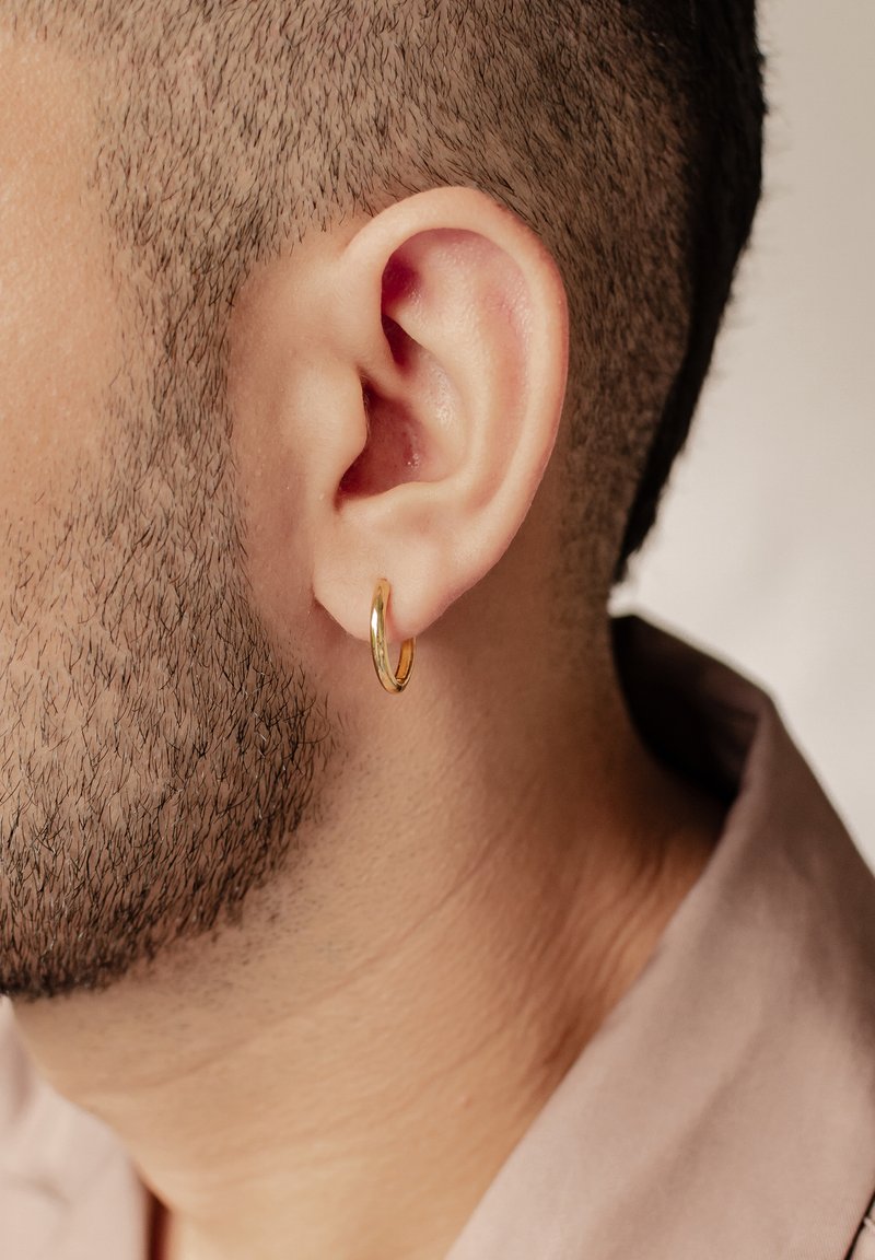 Orecchino a cerchio dorato indossato all'orecchio sinistro, con una forma liscia e arrotondata e una finitura lucida. Primo piano dell'orecchio e dei capelli corti.
