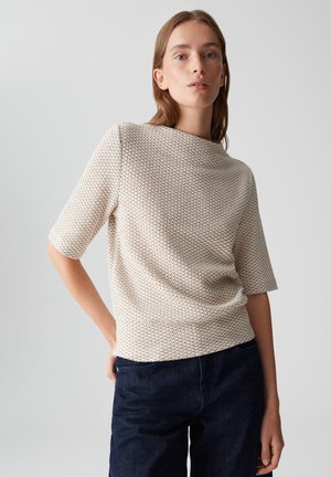 Short-sleeve top in cream with a textured knit pattern. Features a boat neck and is paired with dark blue jeans. Minimalistic design.