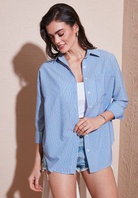 Blue and white striped button-up shirt with long sleeves and chest pocket, made from lightweight fabric, relaxed fit, paired with denim shorts.