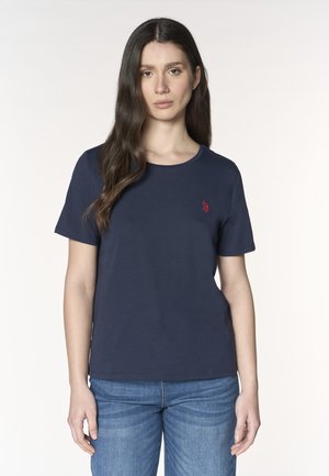 Giovane donna con lunghi capelli scuri che indossa una maglietta blu navy a maniche corte e jeans blu, in piedi davanti a uno sfondo semplice.