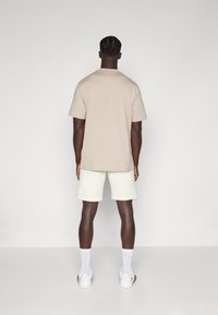 Beige T-Shirt mit Rundhalsausschnitt, lässiger Schnitt, kombiniert mit hellen Cremeshorts. Das Modell trägt weiße Sneakers und kniehohe weiße Socken. Schlichtes Design.