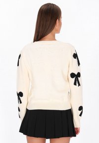 Hosszú barna hajú nő, aki krémszínű pulóvert visel fekete masni mintákkal az ujjakon és fekete félig áttetsző mini szoknyát, háttal áll.