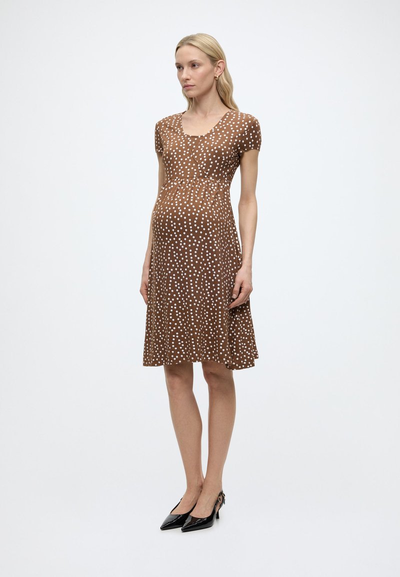 Γυναίκα που φοράει καφέ φόρεμα μέχρι το γόνατο με λευκά πουά και μαύρες γόβες, στέκεται μπροστά σε απλό φωτεινό φόντο.