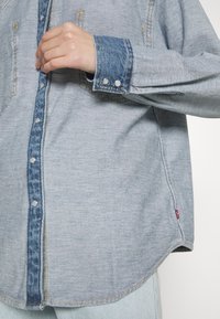 Gros plan sur une chemise en denim bleu clair avec une patte de boutonnage et des poignets bleu foncé, portée par une personne en train de régler les boutons avant.