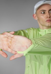 Helle grüne Sportjacke aus leichtem, glattem Material; mit elastischen Armöffnungen und verstellbaren Laschen für einen sicheren Sitz.