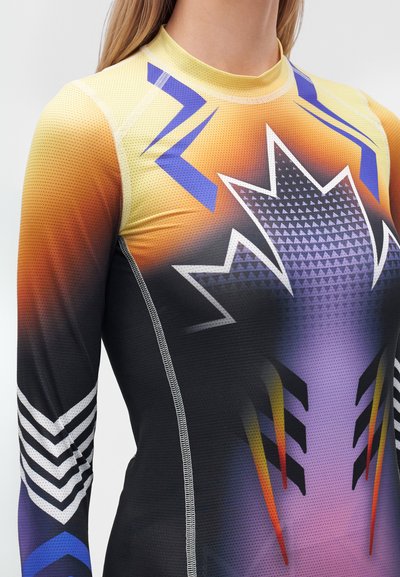 Gros plan sur un haut de sport à manches longues avec un dégradé orange, violet, noir et des motifs géométriques, porté par une personne aux cheveux blonds.