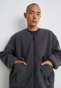 Grå fleecejakke med lynlås foran, oversized fit og kontrasterende mørkegrå stofdetaljer. Har trykknap-lommer i siden.