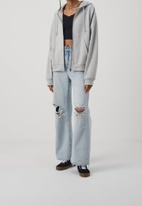 Šedá mikina s oversized střihem a kapucí, spárovaná se světle modrými roztrhanými širokými džíny s velkými dírami a černými teniskami.