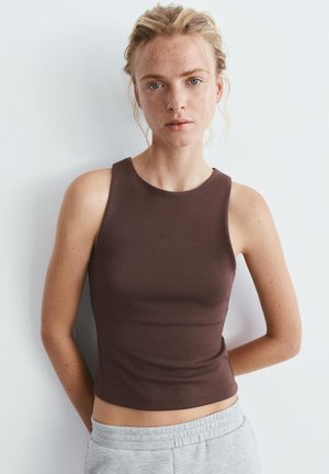 Jeune femme à la peau claire et aux taches de rousseur, portant un haut marron sans manches et un pantalon gris clair, debout devant un fond blanc.