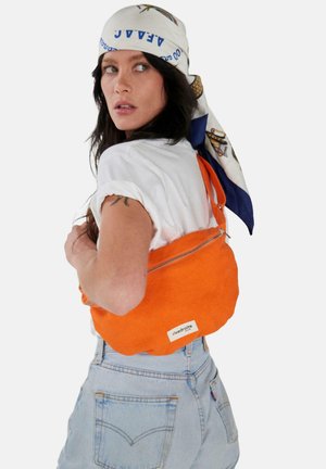 Femme portant un t-shirt blanc, un foulard imprimé sur la tête et un jean bleu clair, tenant un sac à bandoulière orange zippé marqué "rivedroite Paris".
