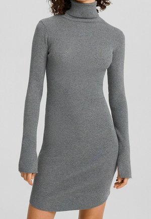 Woman wearing a fitted, long-sleeve, grey ribbed turtleneck dress extending above the knees against a plain background.