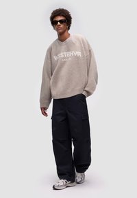 Przesadnie duży beżowy sweter z nadrukiem "WRSTBHVR BERLIN", zestawiony z czarnymi spodniami cargo i metalicznymi sneakersami. Akcesoria to czarne okulary przeciwsłoneczne.