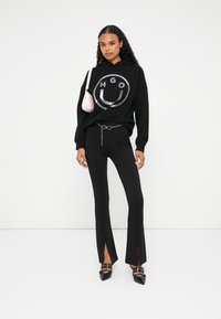 Zwarte hoodie met metalen logo, zwarte flared broek met insnijdingen aan de zoom, en zwarte schoenen met decoratieve gespen. Kleine roze tas.