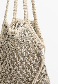 Beige kudottu olkalaukku, jossa on neulottu muotoilu, varustettu tukevilla kahvoilla ja teksturoidulla kuviolla, tummista langoista korostettuna.
