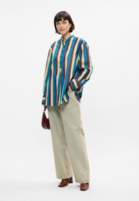 Ριγέ πουκάμισο σε μπλε, μωβ, κίτρινο και λευκό. Χαλαρή εφαρμογή με γιακά και μακριά μανίκια. Συνδυάζεται με μπεζ παντελόνι σε φαρδιά γραμμή και καφέ τακούνια.