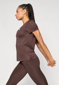 Καφέ αθλητική μπλούζα με κοντά μανίκια, κατασκευασμένη από μαλακό ύφασμα, με πλευρικές σχισμές· συνδυασμένη με καφέ κολάν που ταιριάζουν.