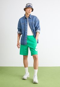 Chemise en denim à boutons, débardeur blanc, shorts verts avec logo blanc, baskets blanches et chapeau seau à carreaux sur fond uni.