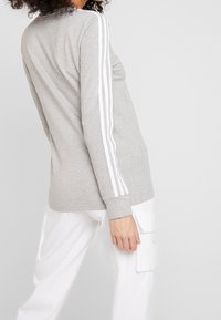 Grå långärmad skjorta i bomull med vita ränder längs axlarna. Har en avslappnad passform och en enkel rund halsringning.