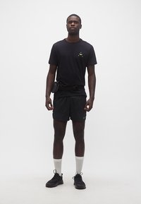 Άνδρας στέκεται φορώντας μαύρο μπλουζάκι, μαύρο αθλητικό σορτς, μαύρα αθλητικά παπούτσια, λευκές κάλτσες και μαύρη τσάντα μέσης σε λευκό φόντο.