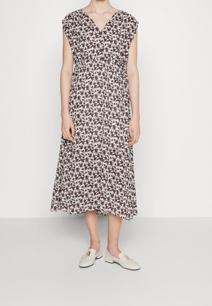 Vrouw die een mouwloze, V-hals, enkellange jurk draagt met een bruin-wit bloemenprint en witte loafers op een effen achtergrond.